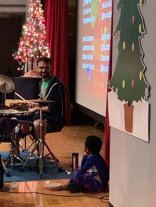 Noah Fuchs plays the drums as student looks on 
